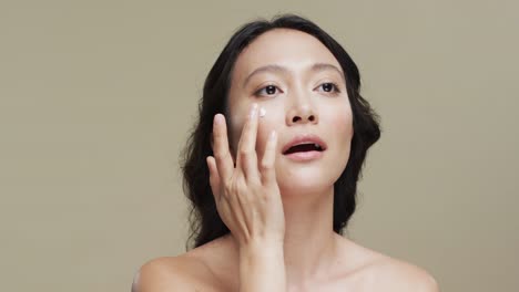 Asian-woman-with-dark-hair-applying-cream-on-her-face-on-beige-background,-copy-space,-slow-motion