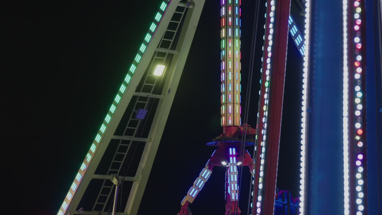 Description: Close-up of a giant pendulum or drop tower ride, illuminated by a spectacular display of colorful, flashing lights against the dark night sky