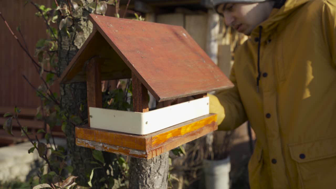Un giovane uomo che indossa una giacca invernale gialla, che controlla la piccola casetta per uccelli su un ramo d'albero - Primo piano