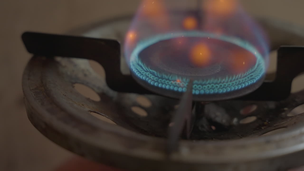 Blå gasflamme danser på brænder, turkis ring gløder, rusten rist i forgrunden, bløde orange pletter, stabil pilotflamme, omgivende køkkenlys, filmisk slow motion-fornemmelse, tekstureret metal, stille hjemligt.