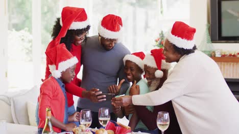 African-american-family-in-santa-hats-using-smartphone-while-standing-near-dining-table-after-having