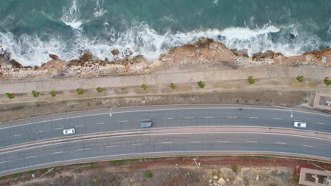 Beautiful-aerial-view-of-cars-passing-by-near-a-bluff-with-water-crashing-on-the-rocks