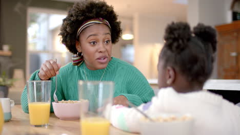 African-american-mother-and-daughter-eating-cereal-with-milk-and-talking-in-kitchen,-slow-motion