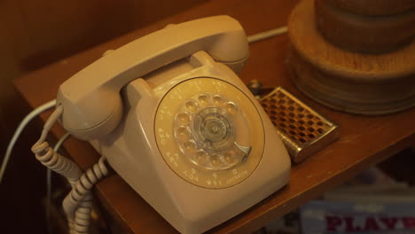 old rotary style white telephone on end table next to curved metal flask by wooden lamp