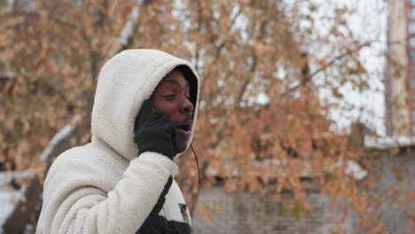 side view of young man in white hoodie conversing over phone with someone, smiling and engaged, blurred background with trees and building