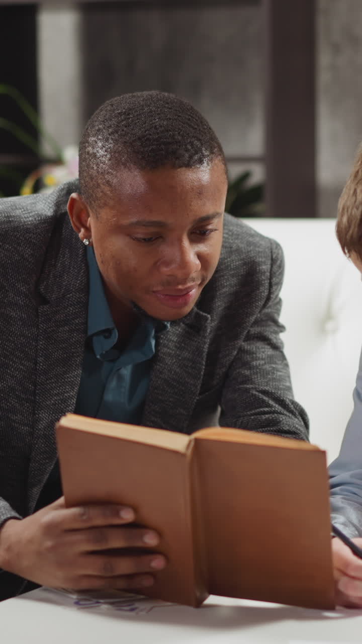 Black private tutor reads book with positive junior schoolboy studying English language at home lesson with native speaker in stylish apartment slow motion