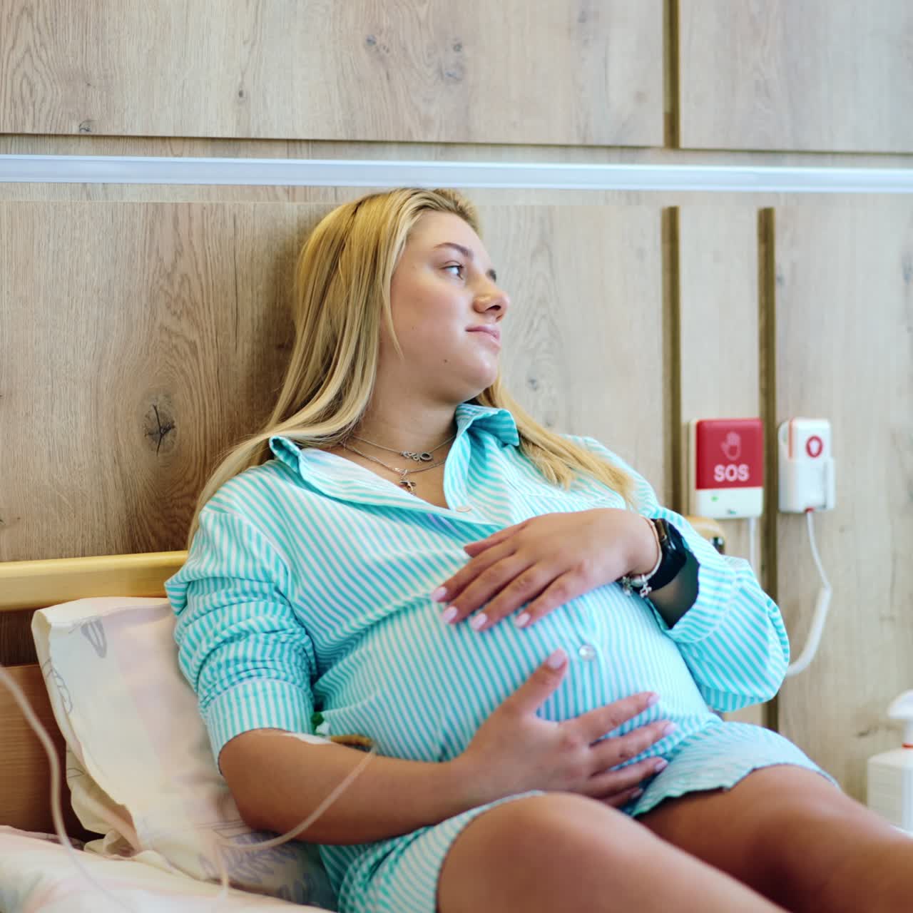 Woman on the last stages of pregnancy lies on bed in hospital. Future mom stroking her big belly while receiving medicine from drop counter