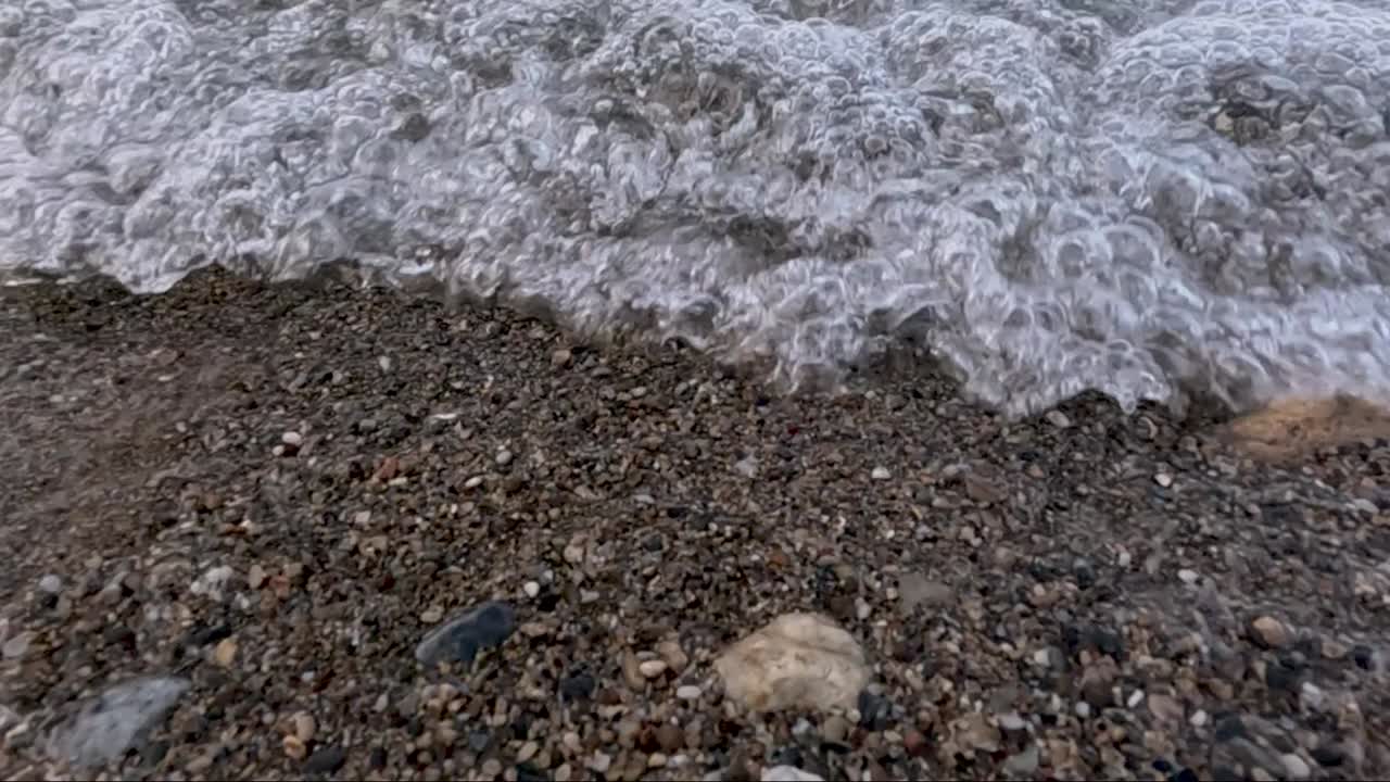 Soft waves wash over a shoreline dotted with vibrant pebbles reflecting sunlight. Tranquility fills the scene as nature's beauty unfolds in this peaceful coastal landscape during the late afternoon