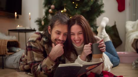 interested guy and girl lying on the floor and reading a christmas book along with a cup of hot drink in a cozy room with a christmas tree and a decorated room in winter evening