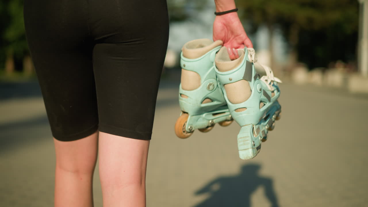 오른손에 시안색 롤러 스케이트를 들고 인 경로에서 야외를 고 있는 여자의 아래 시선, 그녀의 형상의 그림자는 멀리 배경에 초록색과 함께 땅에서 명확하게 볼 수 있습니다.