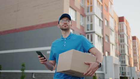 the postman with glasses carries the parcel and looks at the delivery address via mobile phone. search for the address of the delivery customer. delivery guy with a cap and a box