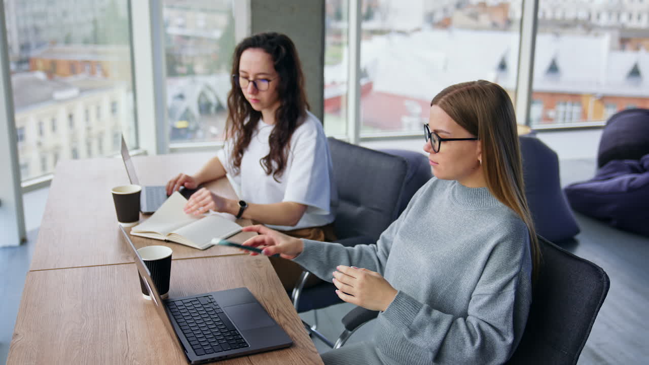 Busy female office employees using laptops. Brunette lady turns the pages of paper book and blonde woman takes a phone.