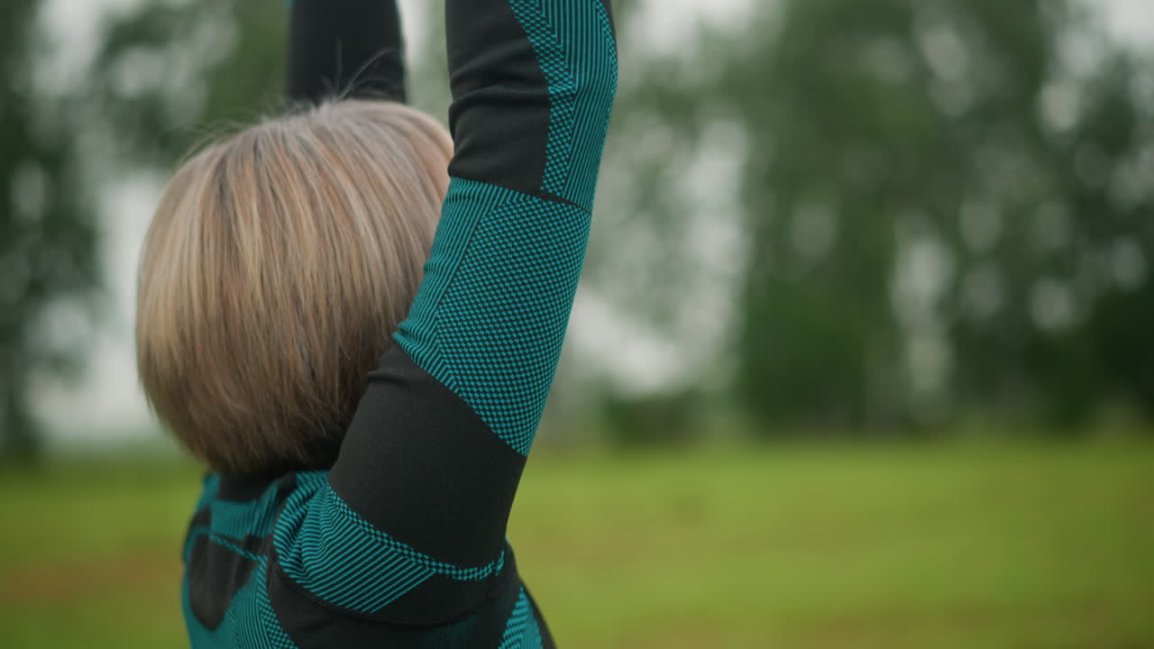 vista lateral de una mujer de mediana edad levantando los brazos y juntando las manos en meditación, practicando la atención plena al aire libre en un campo de niebla cubierto de hierba bajo un cielo nublado