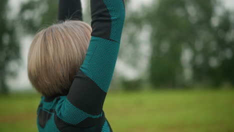 side view of middle-aged woman raising arms and bringing hands together in meditation, practicing mindfulness outdoors in a grassy misty field under a cloudy sky