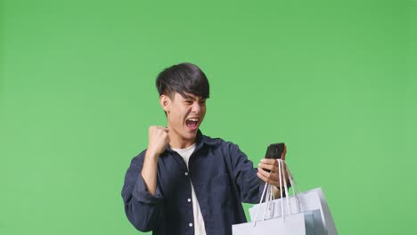 초록색 화면 앞에 서서 휴대 전화 온라인 쇼핑을 사용할 수 있는 행복한 쇼핑 가방을 가진 아시아 쇼핑 남자