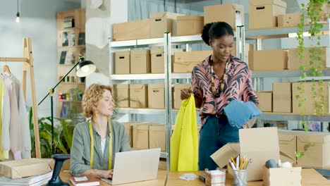 diseñadora afroamericana empacando ropa ordenada en cajas para envío mientras el gerente de ventas caucásico escribe en una laptop en una tienda de ropa de moda