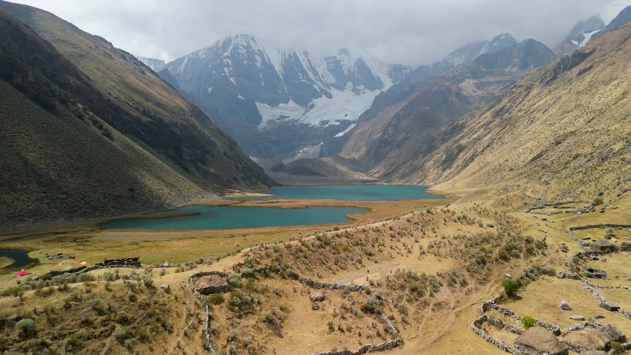 An aerial flyover soars above ancient ruins and a remote campsite, revealing the stunning turquoise glacial lakes at the base of the Huayhuash mountains in Peru