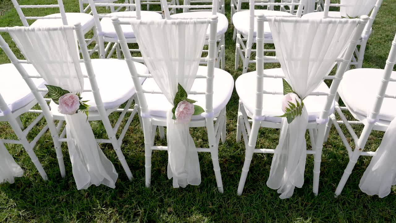 Sedie disposte all'aperto per una cerimonia nuziale. Ogni sedia è rivestita di tessuto bianco e decorazioni floreali. L'erba è verde e il cielo è terso, creando un allestimento semplice per l'evento.