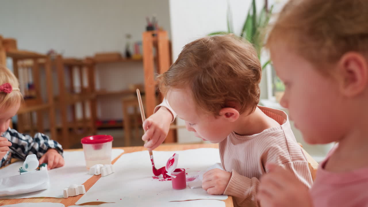 Boy in beige shirt focuses on dipping brush into red paint while carefully painting plaster figurine during classroom art activity, showing creativity and early childhood learning in action