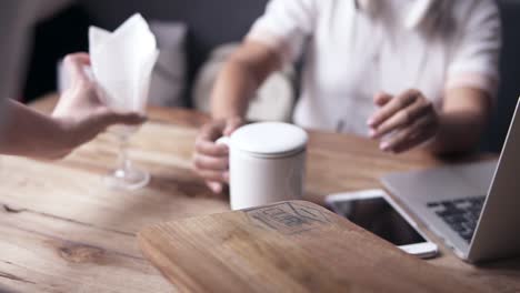 Young-beautiful-woman-in-white,-sitting-in-cafe-on-a-blue-sofa,-waitress-brings-a-cup-of-tea-and-put-it-on-the-table.-There-are-laptop-and-mobile-on-the-table.-White-headphones-on-neck