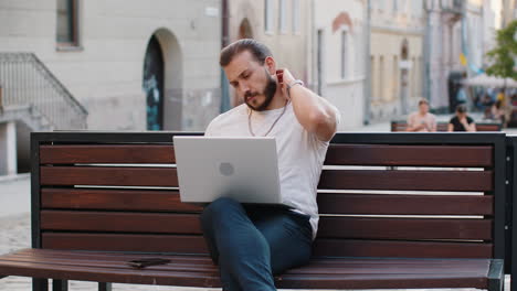 man working online distant job with laptop browsing website chatting sitting on city street bench