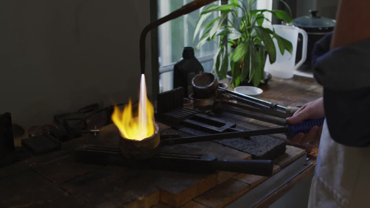 sección media de una joyera caucásica en un taller con un delantal, usando un quemador de gas, derritiendo metal