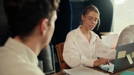 Una-Chica-Rubia-Con-Anteojos-Y-Una-Camisa-Blanca-Se-Comunica-Con-Un-Empleado-Con-Una-Camisa-Blanca,-Sentada-En-Una-Mesa-Con-Una-Computadora-Portátil-En-La-Oficina