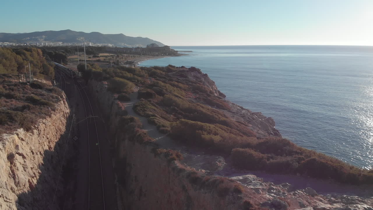 Drone flying over the cliff as train comes out tunnel underneath and drives towards coastal city passes underneath towards