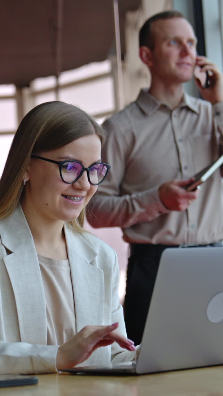 Happy smiling lady works on her laptop at the desk. Man having conversation on the mobile phone at the backdrop. Vertical video