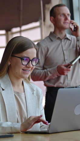 happy smiling lady works on her laptop at the desk. man having conversation on the mobile phone at the backdrop. vertical video