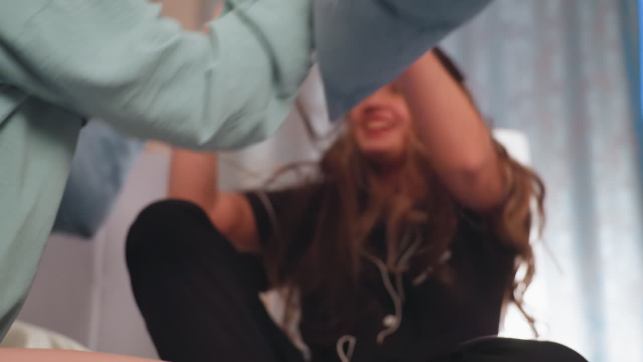 Young girl with long hair covering her face smiles while playfully engaging in pillow fight indoors, her joyful expression and energetic posture capture fun childhood moment