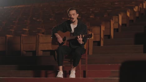 a handsome guy plays an acoustic guitar in an abandoned cinema. the musician sings a song and accompanies on the guitar