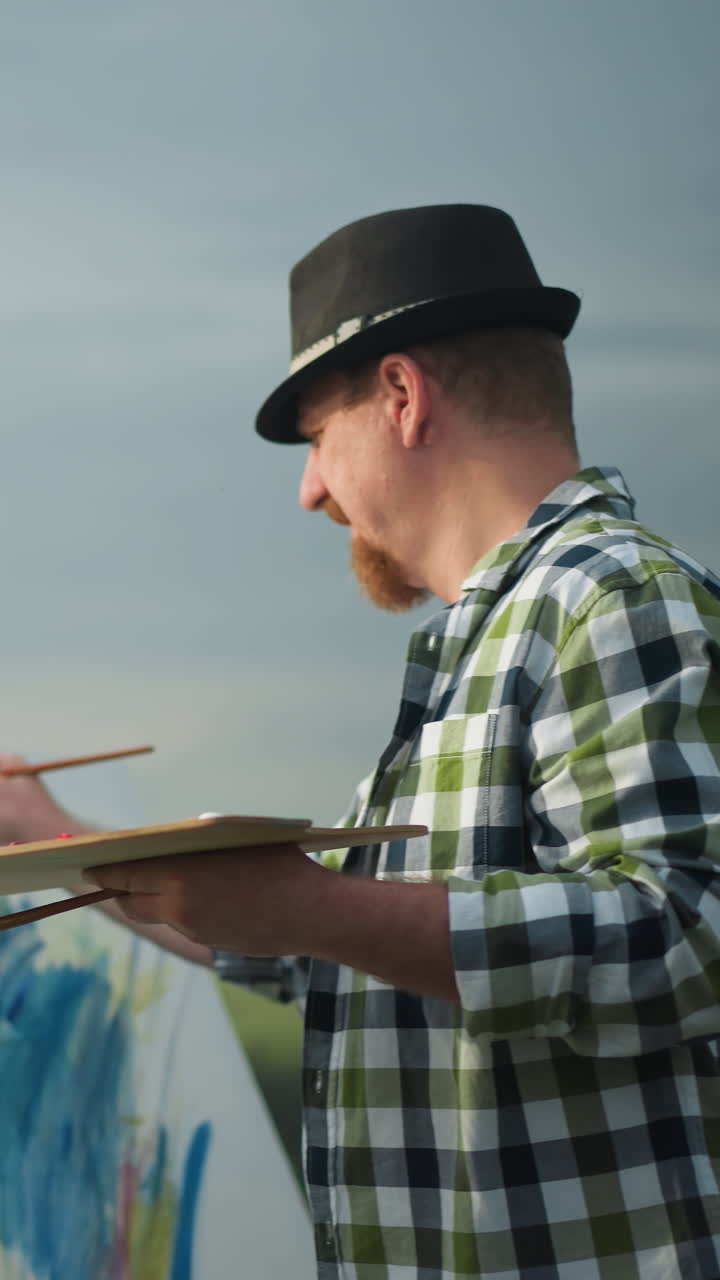 An artist wearing a black hat and a checkered shirt is seen painting on a canvas, holding a palette in a serene grassy environment. A blurred bird is visible flying across the sky