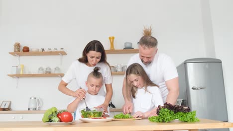 Happy Young Family with two Little Kids Having Fun Cooking Food together in Home Kitchen Happy Young Family with two Little Kids Having Fun Cooking Food together in Home Kitchen Loving Smiling Parents Teaching Little Kids how to Cook Healthy Food Organic Salad or Dinner