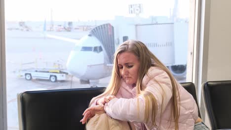A blonde traveler in a pink jacket sits alone in the terminal, lost in thought as she waits for her flight, with the aircraft visible outside