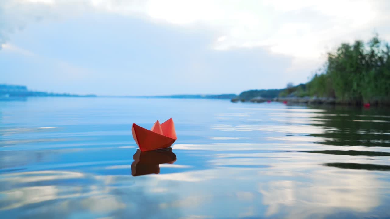 Ship of red paper floating in the blue river at clear day under the blue sky. One paper boat swims on the calm lake in summer