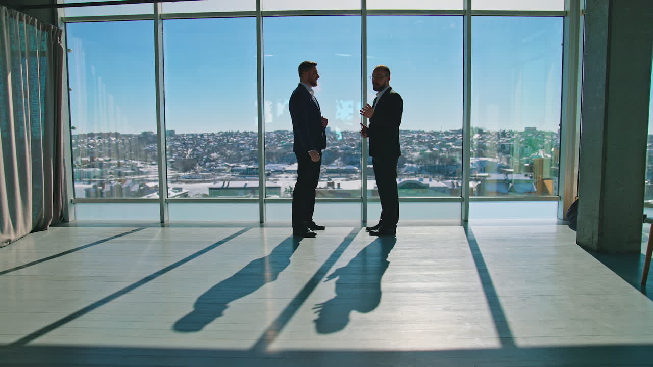 Business partners in office on windows background. Male entrepreneurs in costumes standing near the large window and talking about business plans.