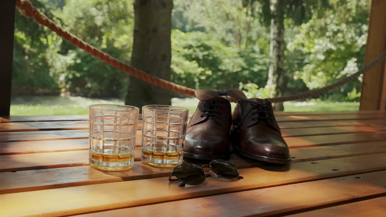 Ripresa a mano di scarpe da sposo, occhiali da sole e due bicchieri nel bosco di giorno, all'aperto