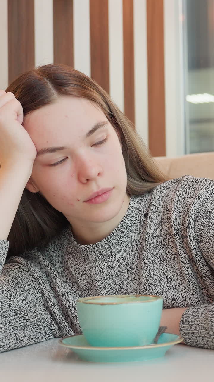 젊은 여성이 카페 테이블에 앉아 청록색 컵으로 커피를 마시고, 스웨터를 입고 스마트폰을 옆에 둔 채, 따뜻한 나무 패널 배경 장면이 바뀌면서 차분한 시선에서 먼 곳을 응시하고 있어요.