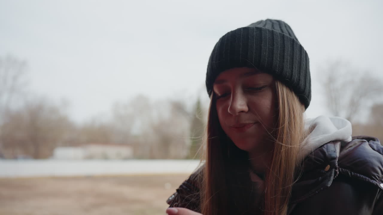 white woman sipping from travel mug at outdoor shelter, brown puffer and knit hat, steaming cup gesture, cold field background, cozy winter ritual, relaxed expression and candid lifestyle scene