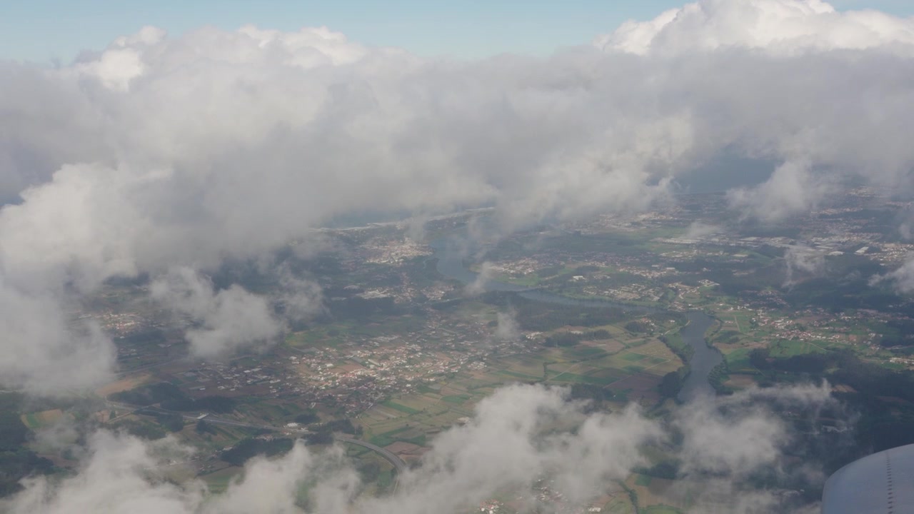 Las nubes cubren el cielo en varias capas. Un avión pasa por encima. Abajo, se ven áreas de tierra con parches de vegetación y pequeños pueblos. La luz del sol ilumina el paisaje.