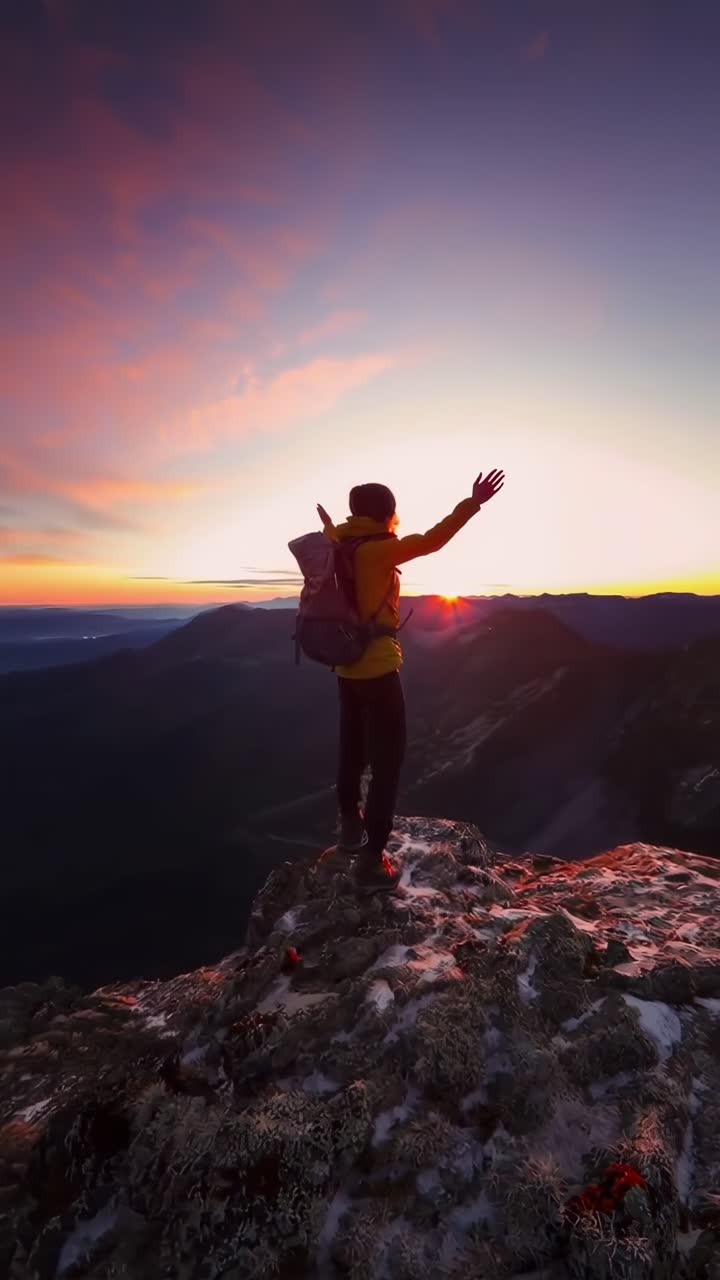 Un escursionista si erge sulla cima di una montagna al tramonto, ripreso da una angolazione bassa, evocando un video cinematografico