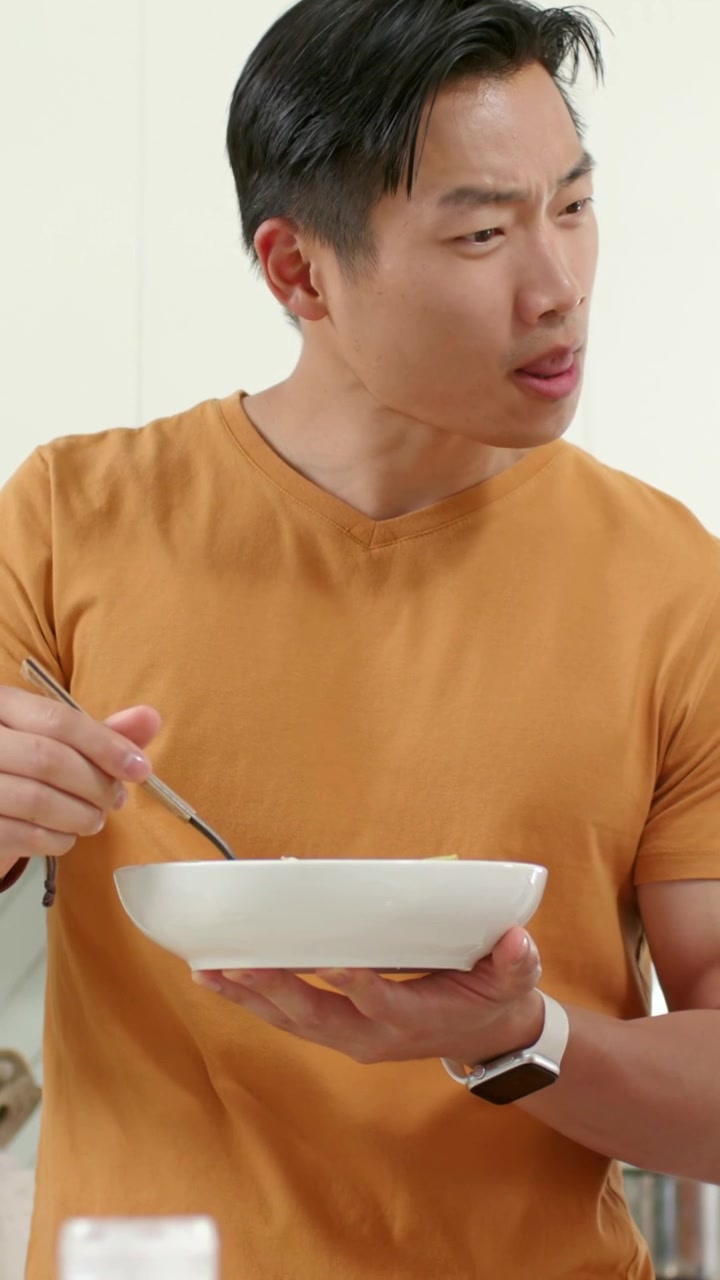 縦型動画：キッチンでオレンジ色のTシャツを着た男性がボウルを持ち、フォークで一口分を持ち上げて料理を味わい評価している。