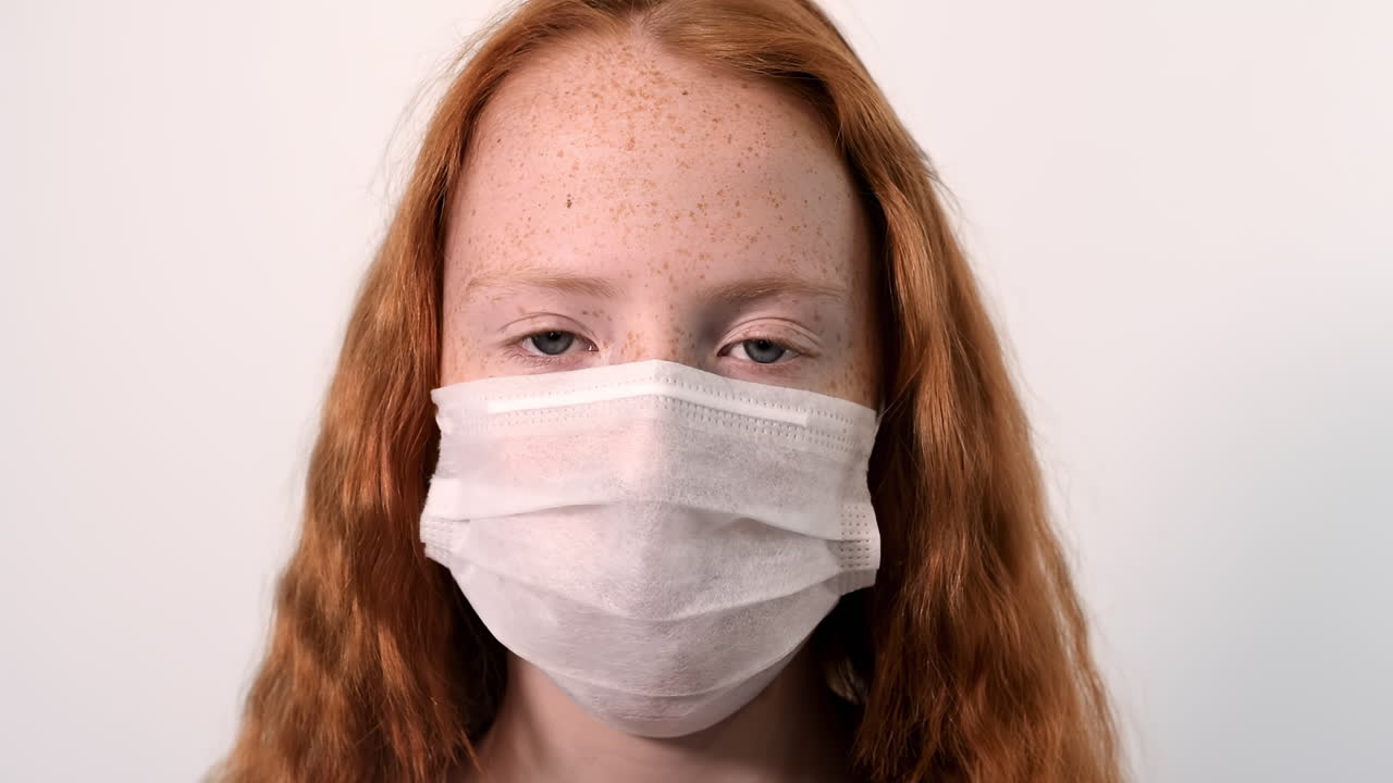 Red haired girl with lots of freckles on the face and blue eyes taking a protective medical mask off the face while looking into the camera. White background. Corona Virus and pandemic idea