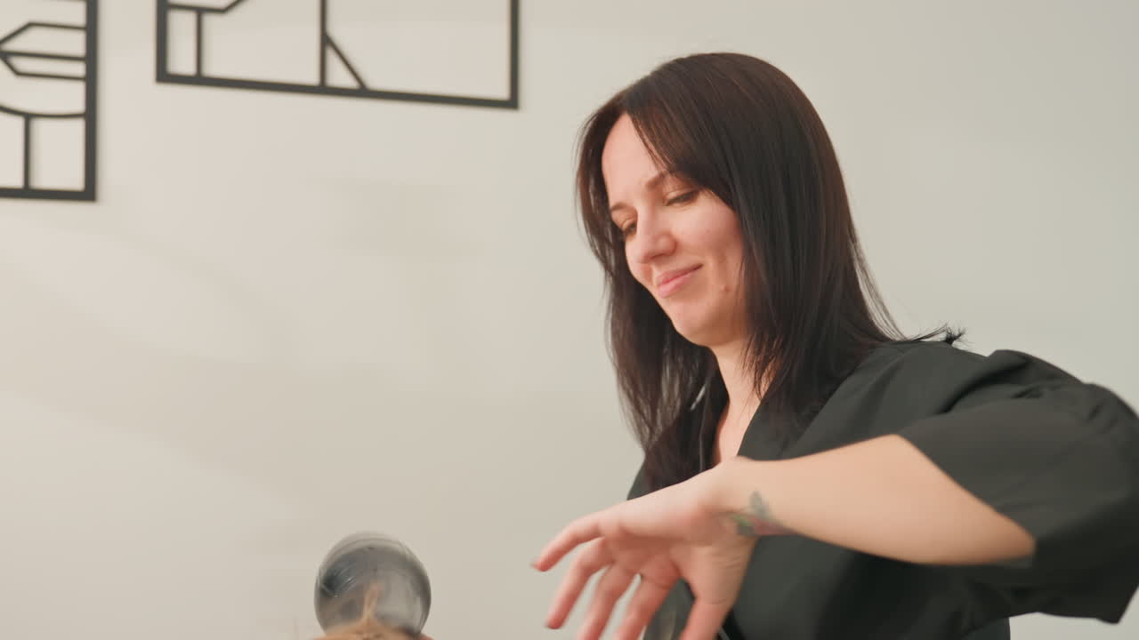 Donna caucasica che si asciuga i capelli con il phon in un luminoso salone domestico, sorridendo mentre mette in piega i capelli scuri di media lunghezza usando un asciugacapelli nero, un vento leggero solleva le ciocche, sfondo con arte murale moderna, umore rilassato e sicuro di sé