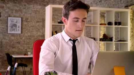 joven empresario guapo está trabajando con una computadora portátil y bebiendo café en la oficina, mirando a la cámara, concepto de negocio, concepto de comunicación