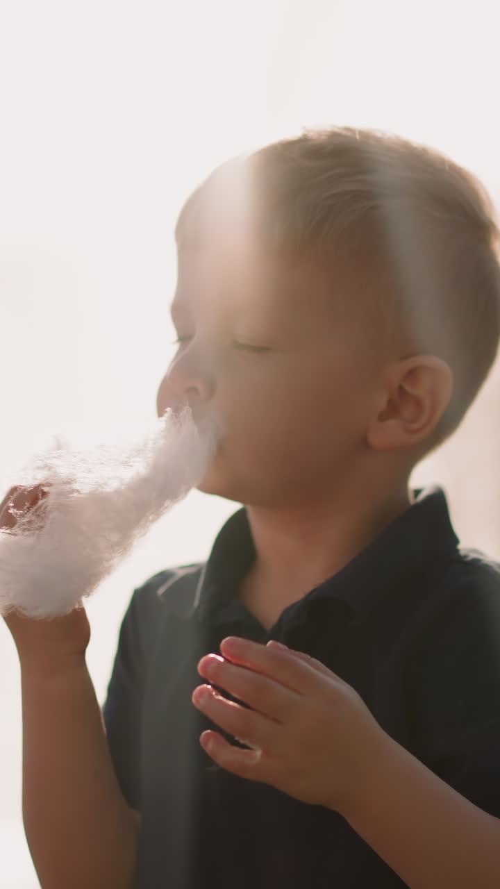 Blond little boy eats large piece of cotton candy against fountain on sunny day closeup slow motion. Happy toddler with dessert. Kid in park