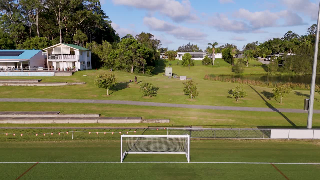 Drone footage captures a soccer field and nearby houses under clear skies, showcasing expansive greenery and serene environment