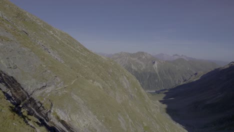 Drone-captures-two-hikers-walking-on-a-trail-along-the-steep-grassy-slope-of-a-mountain-during-the-golden-hour,-showcasing-the-warm-and-enchanting-alpine-landscape