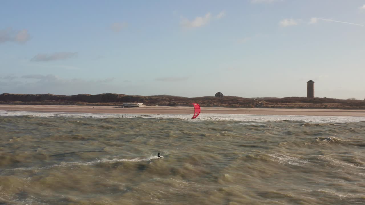 Kitesurfer riding between waves towards a touristic town located behind the dunes. Drone shot